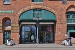 Vorderansicht des roten Backstein-Gebäudes Maritimes Museum Peter Tamm mit grüner Torarkade, Glas-Eingang und zwei historischen Kanonen links und rechts.