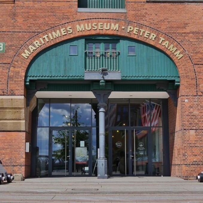Vorderansicht des roten Backsteinbaus Maritimes Museum Peter Tamm mit grünem Bogenportal, Glas-Eingang, US-Flagge innen und zwei Kanonen links und rechts.