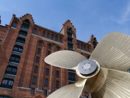 Großer bronzefarbener Schiffsschraubenpropeller im Vordergrund vor einem roten Backsteingebäude des Maritimen Museums unter klarem blauem Himmel.