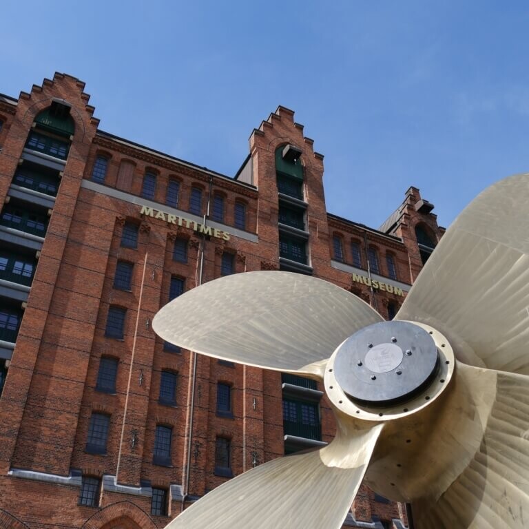 Großer bronzefarbener Schiffsschraubenpropeller im Vordergrund vor einem roten Backsteingebäude des Maritimen Museums unter klarem blauem Himmel.