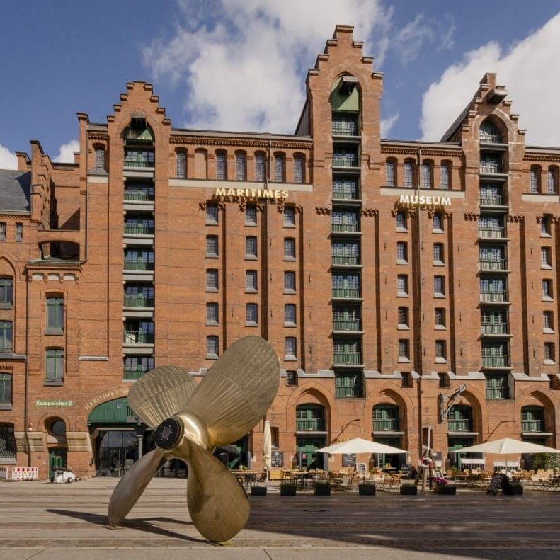 Rotes Backsteingebäude mit Schriftzug Maritimes Museum, davor eine große bronzene Schiffsschraube als Skulptur auf einem Platz mit Cafés und Sonnenschirmen.