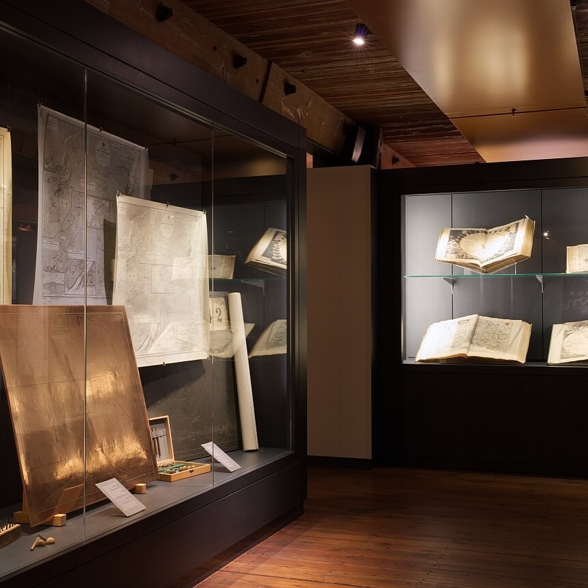 Vitrine mit historischen Seekarten, aufgeschlagenen Atlanten und Plänen hinter Glas in warm beleuchtetem Museumsraum mit Holzfußboden und Decke.