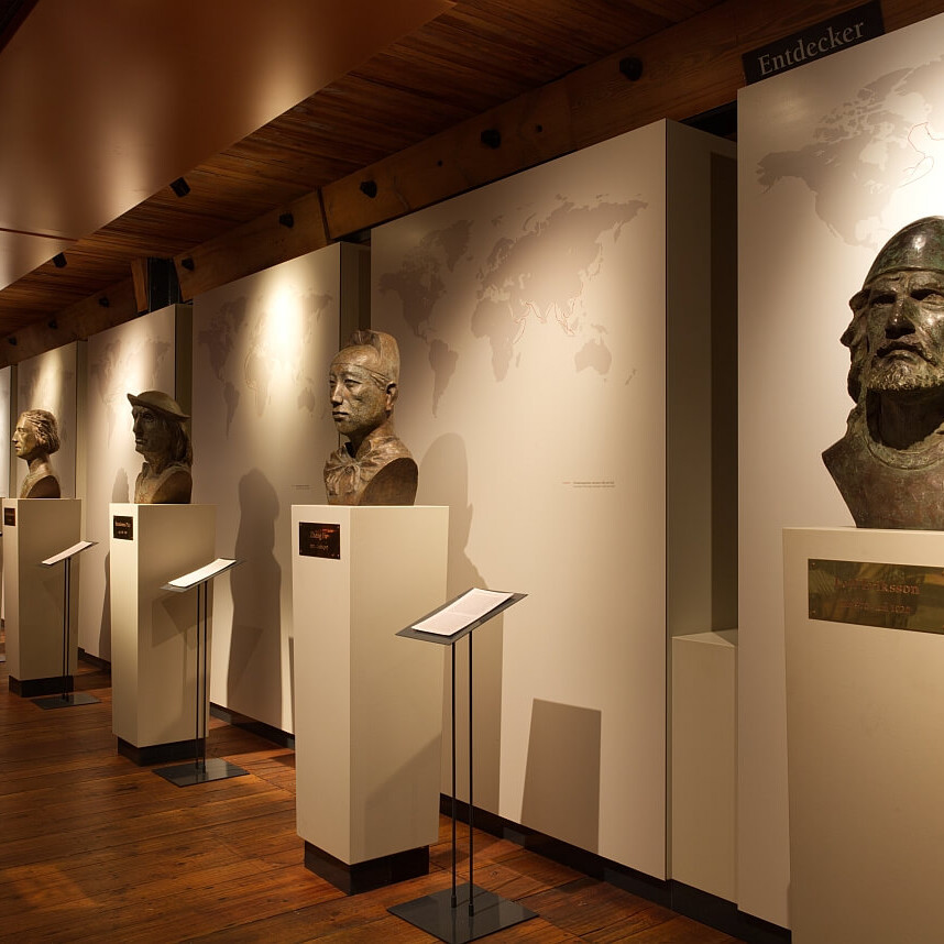 Reihe bronzener Büsten auf weißen Podesten in einem warm beleuchteten Museumsflur mit Holzfußboden und -decke, Weltkarten an der Wand und kleinen Infotafeln.
