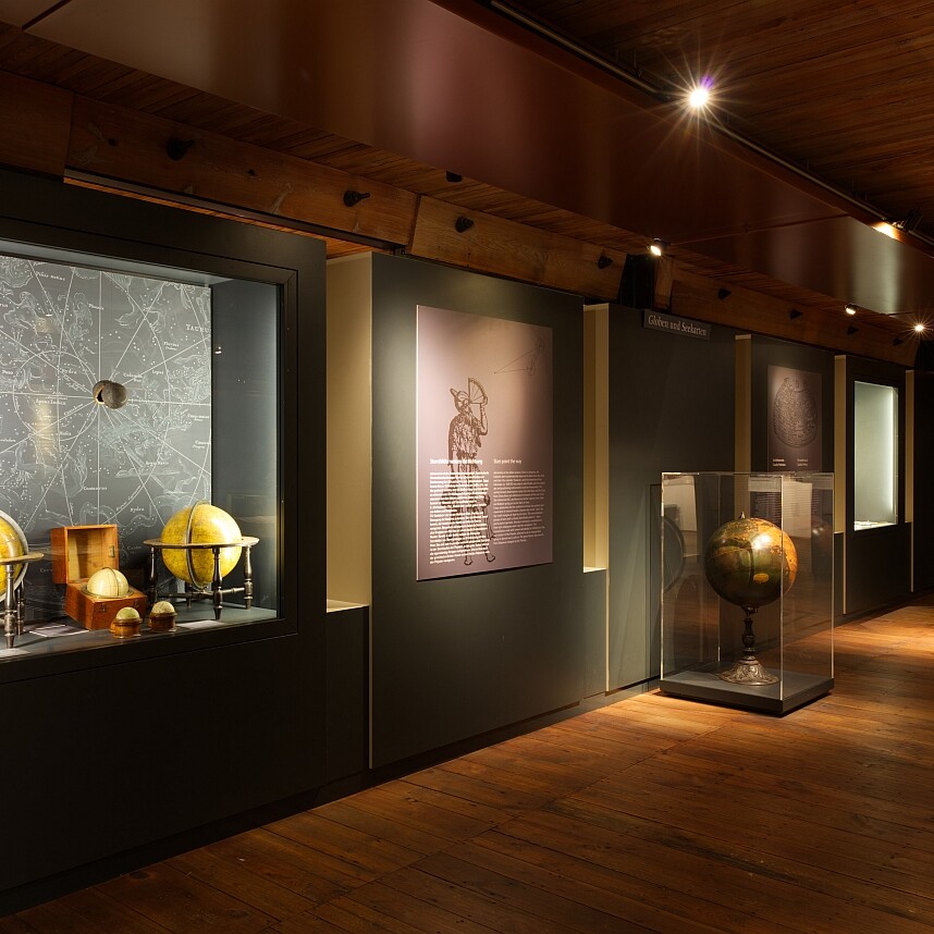 Ausstellungsraum mit Holzdecke und -boden, warm beleuchteten Vitrinen mit historischen Erd- und Himmelsgloben vor einer Sternenkarte und erklärenden Wandtafeln.