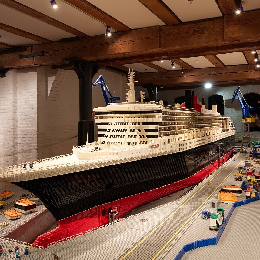 Großes, detailliertes LEGO-Modell eines rot-schwarzen Cunard-Ozeandampfers im Trockendock mit Kränen, Miniaturfahrzeugen und Bauarbeitern in einer Museumshalle.