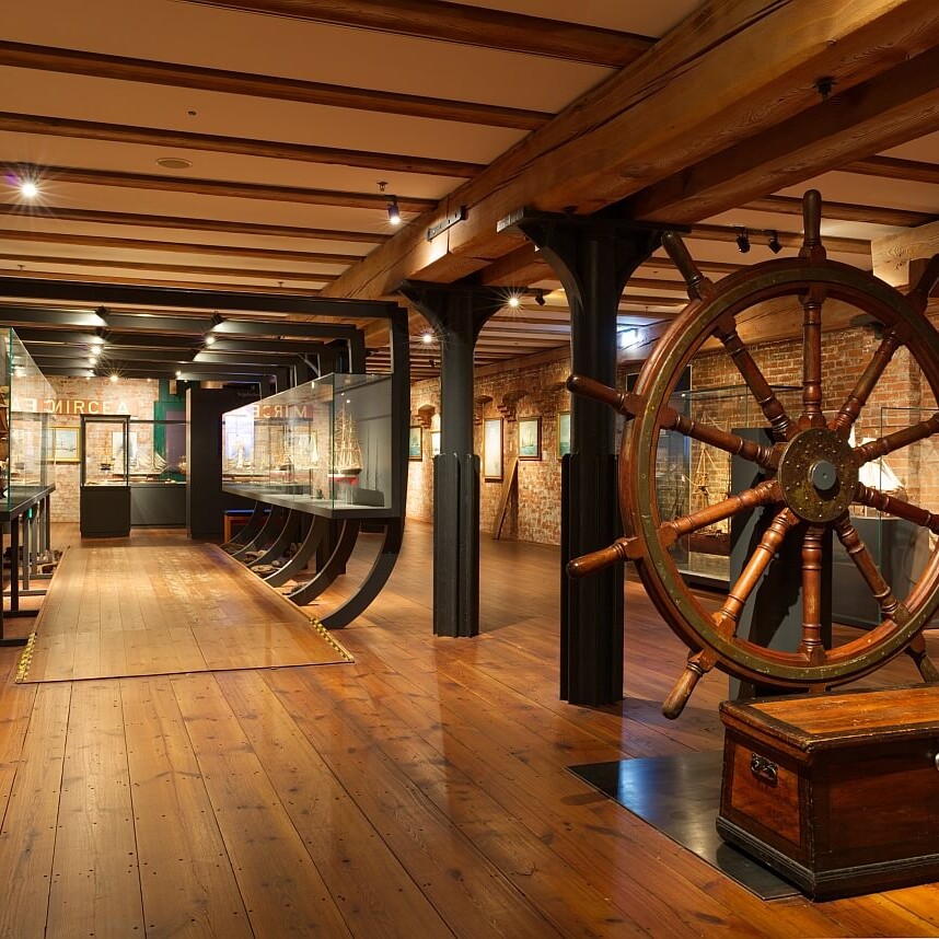 Innenansicht eines maritimen Museums mit warm beleuchtetem Holzboden und Gebälk, großem hölzernem Schiffssteuer im Vordergrund und Modellschiffen in Vitrinen.