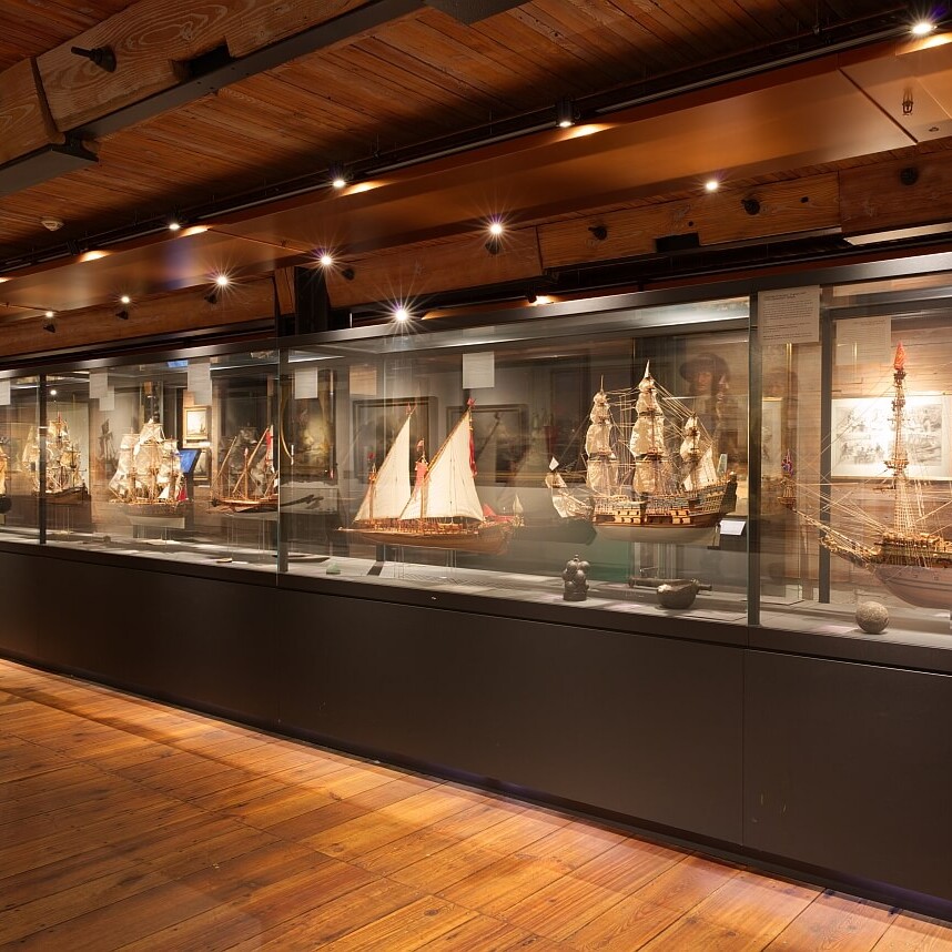 Lange Museumsvitrine mit historischen, fein detaillierten Schiffsmodellen hinter Glas, warm beleuchteter Holzraum mit sichtbaren Deckenbalken und Holzboden.