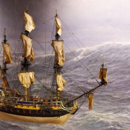 Detailliertes Modell eines dreimastigen Segelschiffs mit gebräunten Segeln und verziertem Rumpf vor aufgewühlter graublauer See.