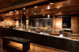 Dunkles Holzmuseum mit warmen Scheinwerfern, Glasvitrinen auf schwarzem Podest zeigen historische Schiffsmodelle; Vordergrund Sitzbank, W&auml;nde mit Bildern.