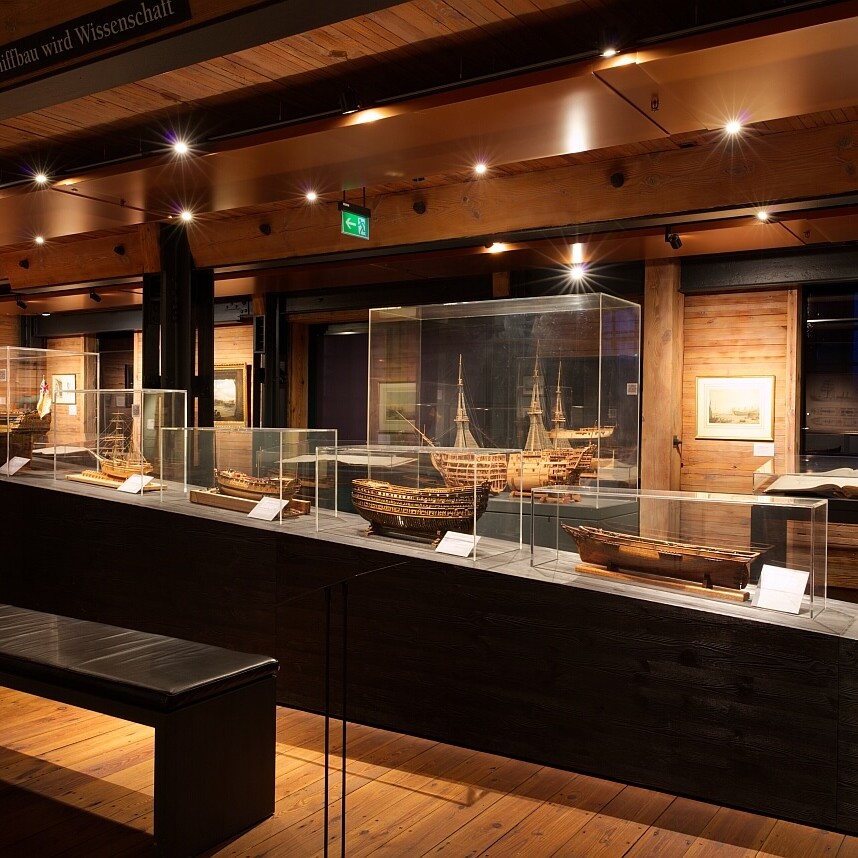 Dunkles Holzmuseum mit warmen Scheinwerfern, Glasvitrinen auf schwarzem Podest zeigen historische Schiffsmodelle; Vordergrund Sitzbank, Wände mit Bildern.