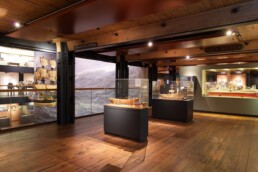 Museumshalle mit Holzfu&szlig;boden und Balkendecke, Glasvitrinen mit Schiffsmodellen, gro&szlig;es Meereswandbild im Hintergrund und warme, ged&auml;mpfte Beleuchtung.