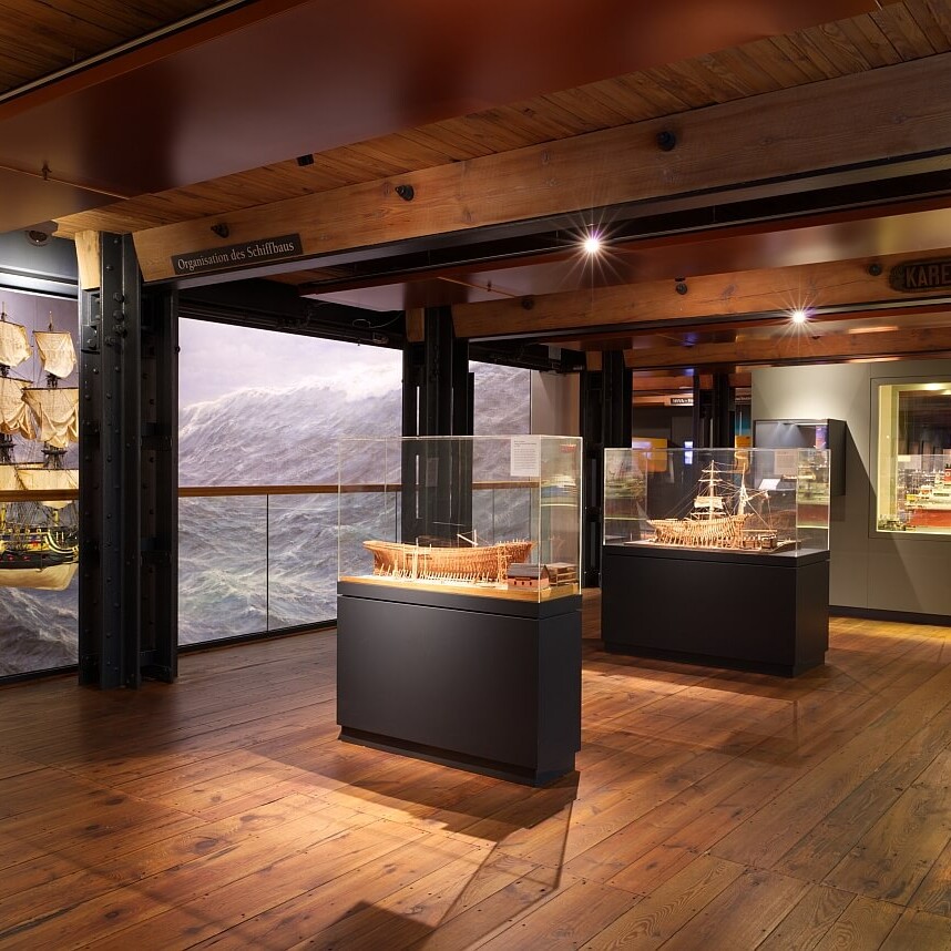 Museumshalle mit Holzfußboden und Balkendecke, Glasvitrinen mit Schiffsmodellen, großes Meereswandbild im Hintergrund und warme, gedämpfte Beleuchtung.