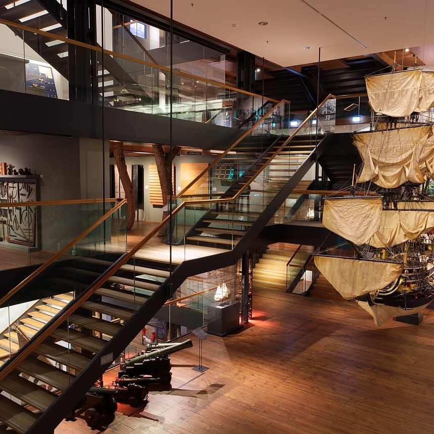 Großer Museumsraum mit hängendem Segelschiffsmodell, mehrstöckigen Glas- und Holztreppen, Holzfußboden, historischen Kanonen und Ausstellungsstücken.