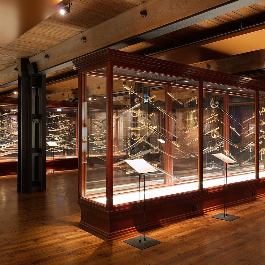 Weitläufige Museumshalle mit warmem Licht und Holzboden, große Glasvitrinen, darin zahlreiche historische Säbel, Degen und Paradewaffen mit goldenen Griffen.