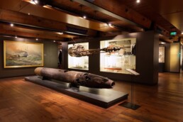 Museumsraum mit verrostetem Torpedo auf Podest, hängendem Antrieb, Vitrinen mit Schiffsmodellen und Seeschlachtgemälde an der Wand.