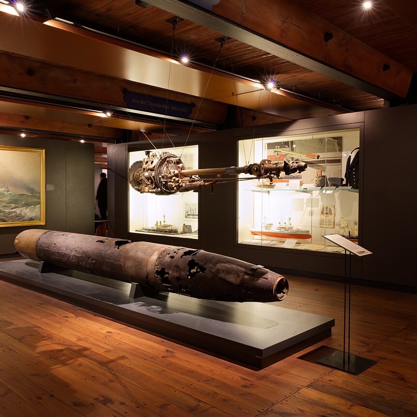 Museumsraum mit verrostetem Torpedo auf Podest, hängendem Antrieb, Vitrinen mit Schiffsmodellen und Seeschlachtgemälde an der Wand.
