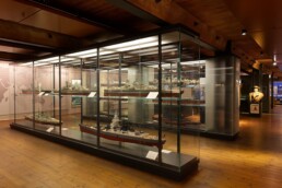 Museumsausstellung mit beleuchteten Glasvitrinen, in denen Schiffsmodelle stehen; warmer Holzboden und Deckenbalken, Büste im Hintergrund.