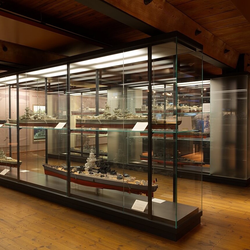 Museumsausstellung mit beleuchteten Glasvitrinen, in denen Schiffsmodelle stehen; warmer Holzboden und Deckenbalken, Büste im Hintergrund.