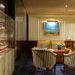 Gemütliche Kabinen- oder Wohnzimmerszene mit Vitrinen-Modellschiff links, zwei blau-gelb gestreiften Polstersesseln am runden Holztisch, Sofa, Ölgemälde und warmem Licht.