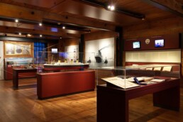 Warm beleuchteter Ausstellungsraum mit sichtbaren Holzbalken und Boden, Vitrinen mit Schiffsmodellen, Dokumenten, historischen Fotografien und Infotafeln.