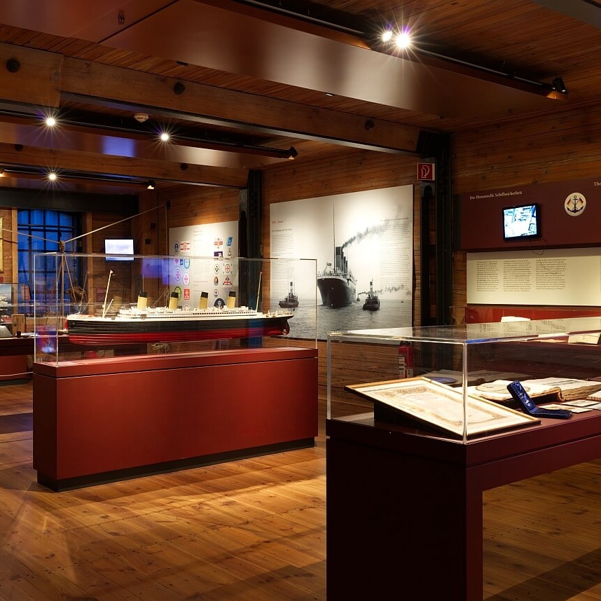 Warm beleuchteter Ausstellungsraum mit sichtbaren Holzbalken und Boden, Vitrinen mit Schiffsmodellen, Dokumenten, historischen Fotografien und Infotafeln.
