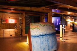 Warm beleuchteter Museumsraum mit Holzdecken, im Vordergrund ein gro&szlig;formatiges blau-gr&uuml;nes Relief des Meeresbodens und der K&uuml;stenlinie, umgeben von Messinstrumenten.