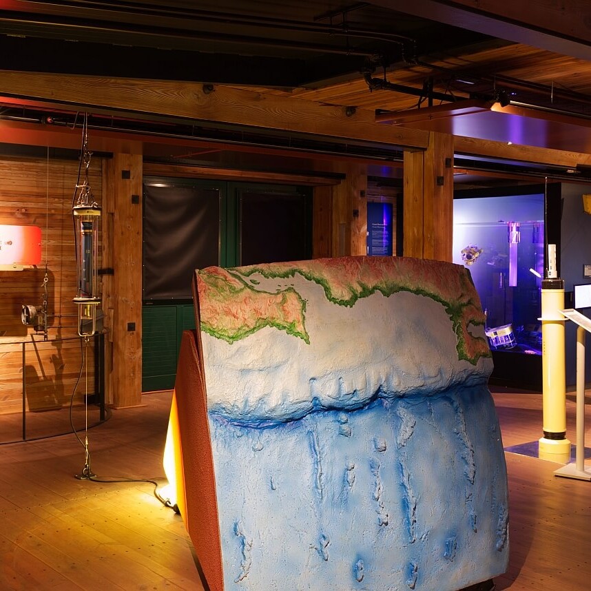 Warm beleuchteter Museumsraum mit Holzdecken, im Vordergrund ein großformatiges blau-grünes Relief des Meeresbodens und der Küstenlinie, umgeben von Messinstrumenten.