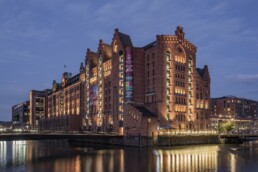 Beleuchtetes historisches rotbraunes Backsteingebäude im Lagerhausstil am Wasser, mit Lichterspiegelung auf der Oberfläche