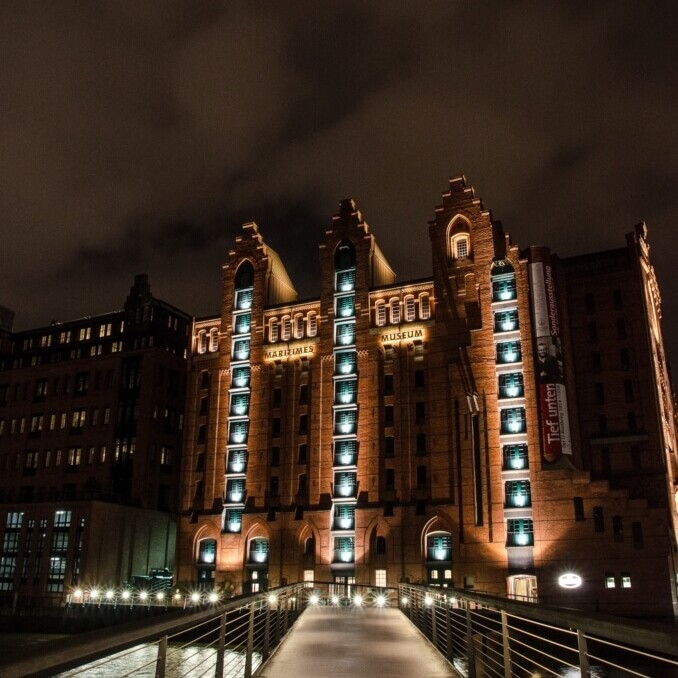 Ein beleuchteter Fußgängersteg führt über einen Kanal zu einem hohen roten Backsteingebäude mit turmartigen Erkern und vertikalen Lichtstreifen bei Nacht.