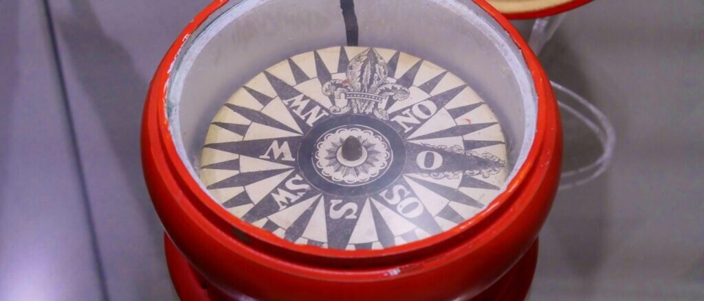 Roter Kompassbehälter mit sichtbarer Kompassrose und markierten Himmelsrichtungen unter Glas.