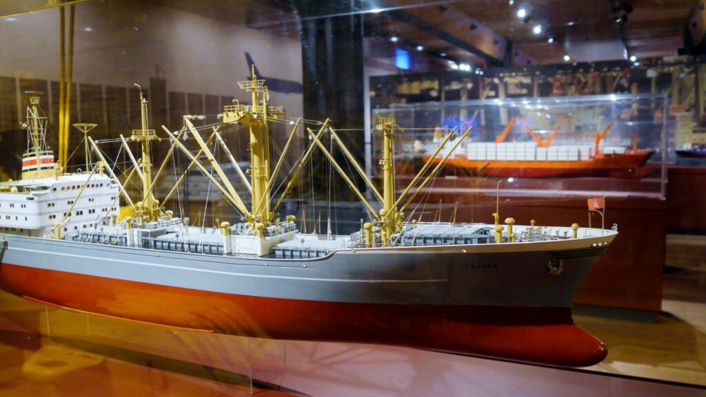 Detailliertes Schiffsmodell mit grauem Rumpf, rotem Unterwasserteil und gelben Ladekränen in einer Museumsvitrine.