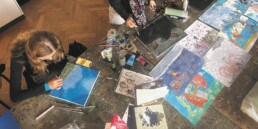 Draufsicht auf einen großen, fleckigen Arbeitstisch voller Pinsel, Farbtuben, Stifte und ausgelegter Zeichnungen, an dem zwei Personen sitzen und malen — links wird an einer blauen Meeresleinwand gearbeitet, rechts an dunkleren und farbigen Illustrationen.
