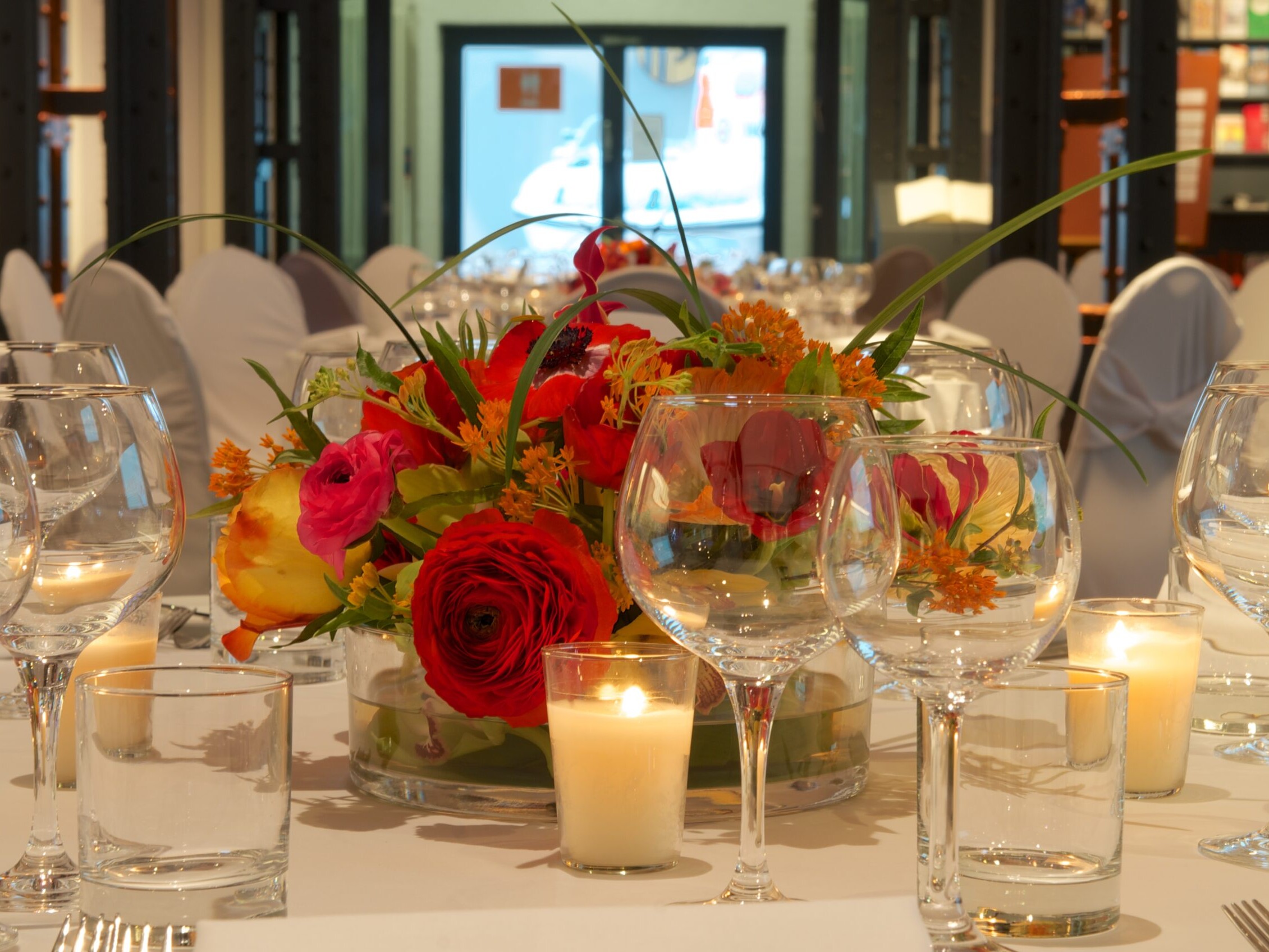 Festlich gedeckter Tisch mit buntem Blumenarrangement, brennenden Kerzen und vielen Weingläsern.