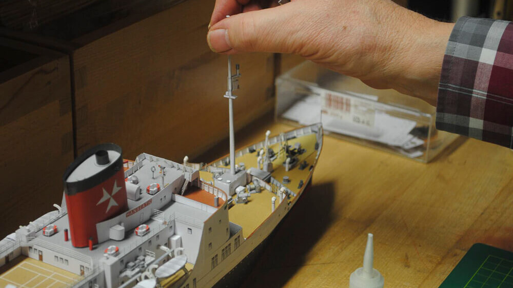 Hand mit Pinzette montiert Mast an detailliertem Modellschiff auf Werkbank, sichtbar Schornstein mit rotem Stern, Kleberflasche und Schneidematte daneben.