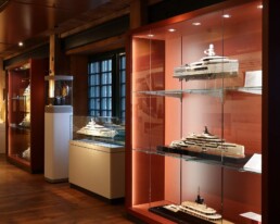 Beleuchteter Ausstellungsraum mit warmem Holzfu&szlig;boden und -decke, in dem hinter Glasvitrinen mehrere detaillierte Schiffs- und Yachtmodelle vor rot-orangem Hintergrund pr&auml;sentiert werden.