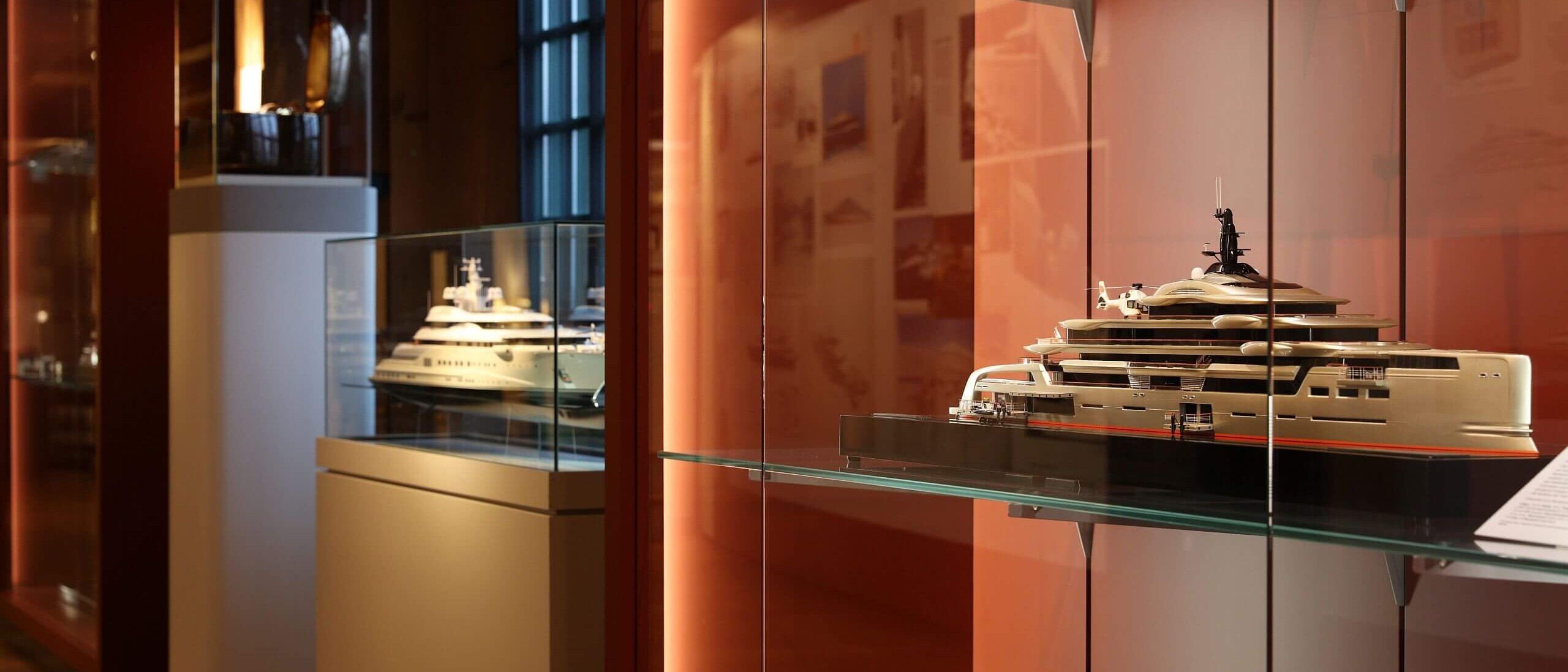 Modell einer modernen Luxus‑Yacht in einer Glasvitrine auf einem Glasregal in einem warm beleuchteten Museumssaal mit roten Wänden, daneben weitere Schiffsmodelle in Vitrinen.
