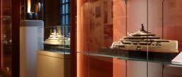 Modell einer modernen Luxus‑Yacht in einer Glasvitrine auf einem Glasregal in einem warm beleuchteten Museumssaal mit roten W&auml;nden, daneben weitere Schiffsmodelle in Vitrinen.