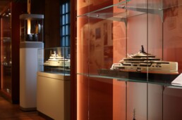 Modell einer modernen Luxus‑Yacht in einer Glasvitrine auf einem Glasregal in einem warm beleuchteten Museumssaal mit roten Wänden, daneben weitere Schiffsmodelle in Vitrinen.