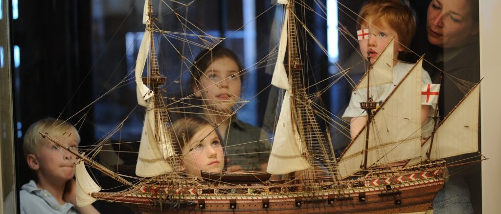 Kinder und Frau betrachten ein detailliertes Segelschiffsmodell mit Takelage und wei&szlig;en Segeln hinter einer Vitrine.