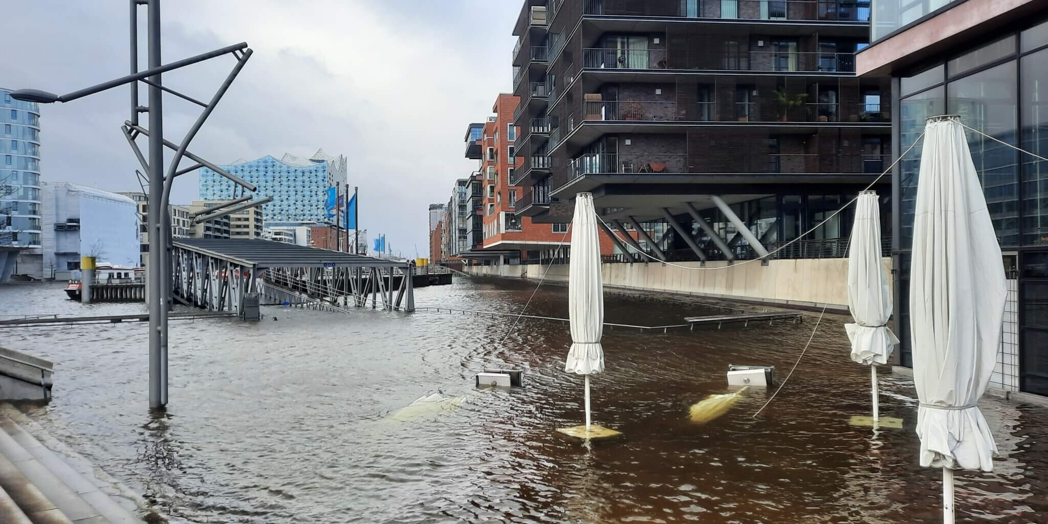 Überfluteter Hafen mit teilweise unter Wasser stehenden, gebundenen Sonnenschirmen, moderner Wohnbebauung und der Elbphilharmonie unter grauem Himmel.