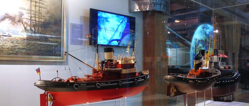 Zwei detaillierte Schlepper-Modelle in einer Museumsvitrine mit roten und schwarzen R&uuml;mpfen, am Heck des linken Modells eine deutsche Flagge; im Hintergrund ein maritimes Gem&auml;lde, ein blauer Monitor und Spiegelungen auf der Glasoberfl&auml;che.