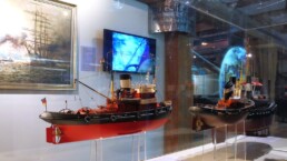 Zwei detaillierte Schlepper-Modelle in einer Museumsvitrine mit roten und schwarzen Rümpfen, am Heck des linken Modells eine deutsche Flagge; im Hintergrund ein maritimes Gemälde, ein blauer Monitor und Spiegelungen auf der Glasoberfläche.