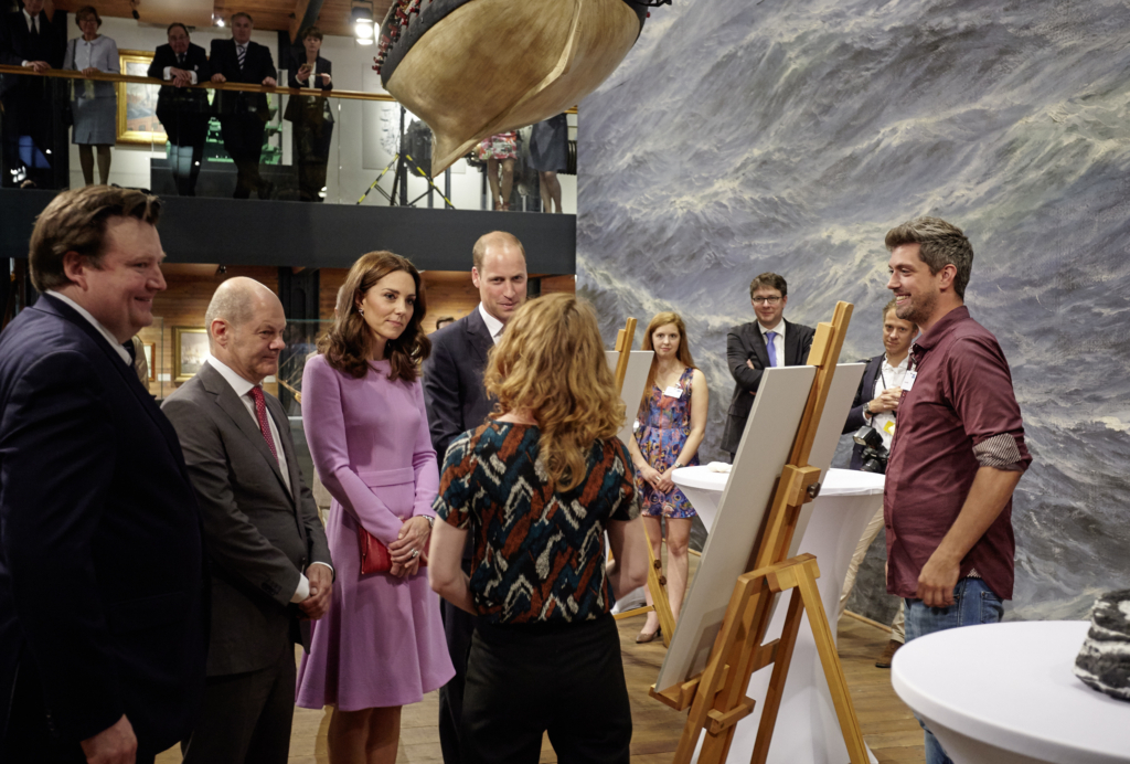 Gruppe gut gekleideter Besucher in einer Ausstellung vor Staffeleien und gro&szlig;em Meeresgem&auml;lde; ein K&uuml;nstler erkl&auml;rt l&auml;chelnd, ein Boot h&auml;ngt dar&uuml;ber.