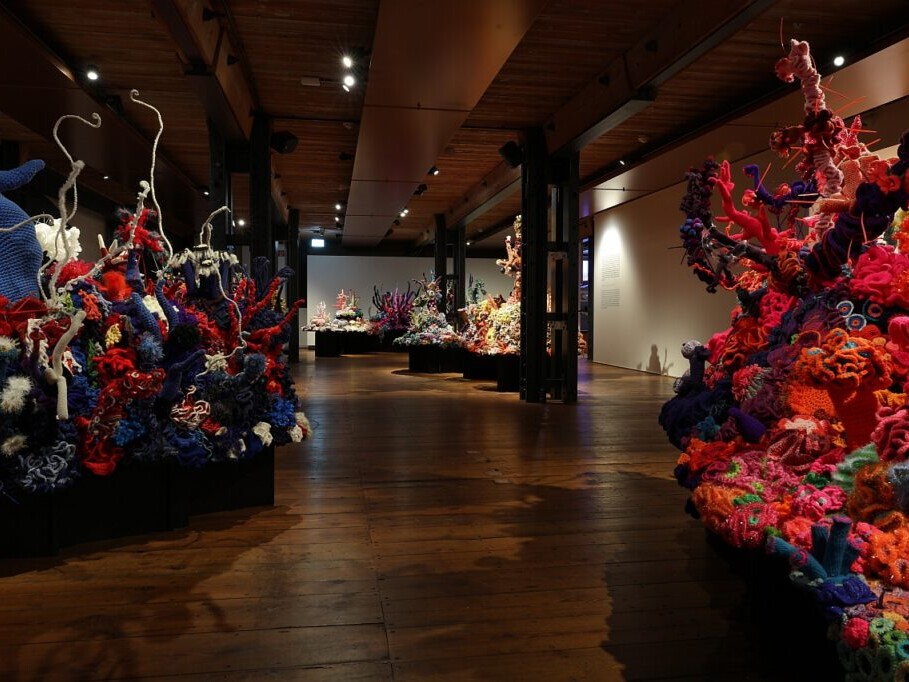 Blick in eine dunkle Galerie mit großformatigen, bunten Häkelinstallationen, die wie Korallenriffe in Rot-, Blau- und Orangetönen auf Podesten wachsen.