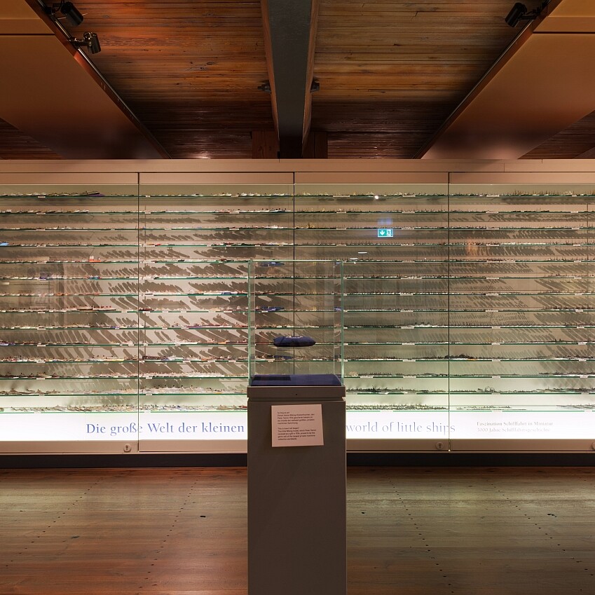 Breite Museumsvitrine vor Holzdecke und -boden, gefüllt mit hunderten Miniaturschiffen auf vielen Glasregalen; mittig auf Sockel ein einzelnes Modell in Glas.