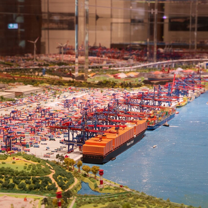 Hafenmodell mit großen Containerfrachtern am Kai, gestapelten bunten Containern, roten Kranen entlang der Mole, blauem Wasser und Windrädern im Hintergrund.
