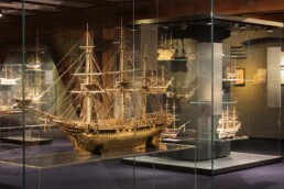 Detailliertes Holzmodell eines Dreimast-Segelschiffs mit feiner Takelage in einer beleuchteten Glasvitrine, umgeben von weiteren Modellschiffen im Museum.