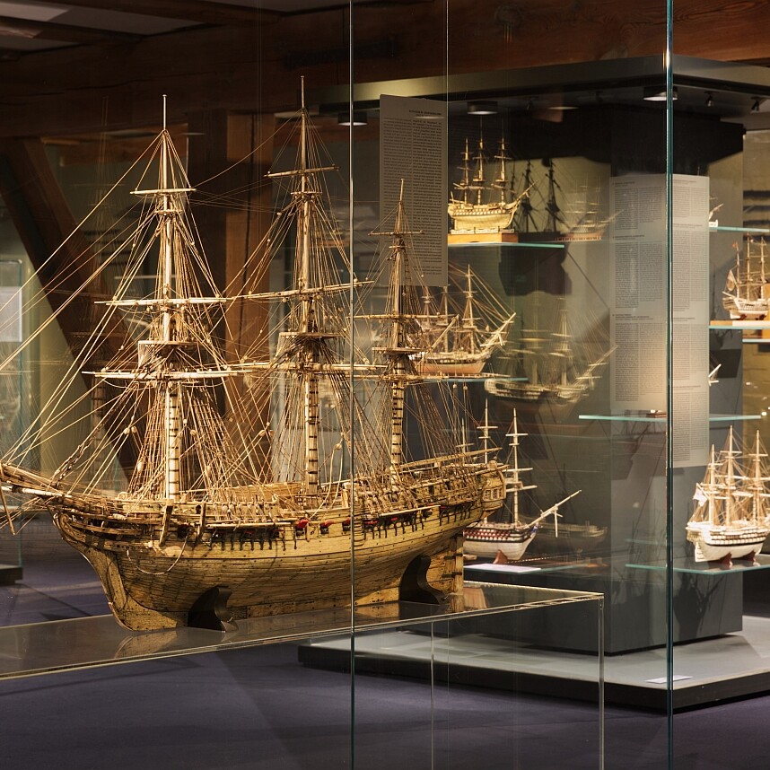 Detailliertes Holzmodell eines Dreimast-Segelschiffs mit feiner Takelage in einer beleuchteten Glasvitrine, umgeben von weiteren Modellschiffen im Museum.