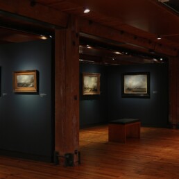 Dimmer Museumsraum mit dunklen Wänden und hölzernem Boden, mehrere gerahmte maritime Gemälde an der Wand, schwere Holzbalken und eine Sitzbank mittig.