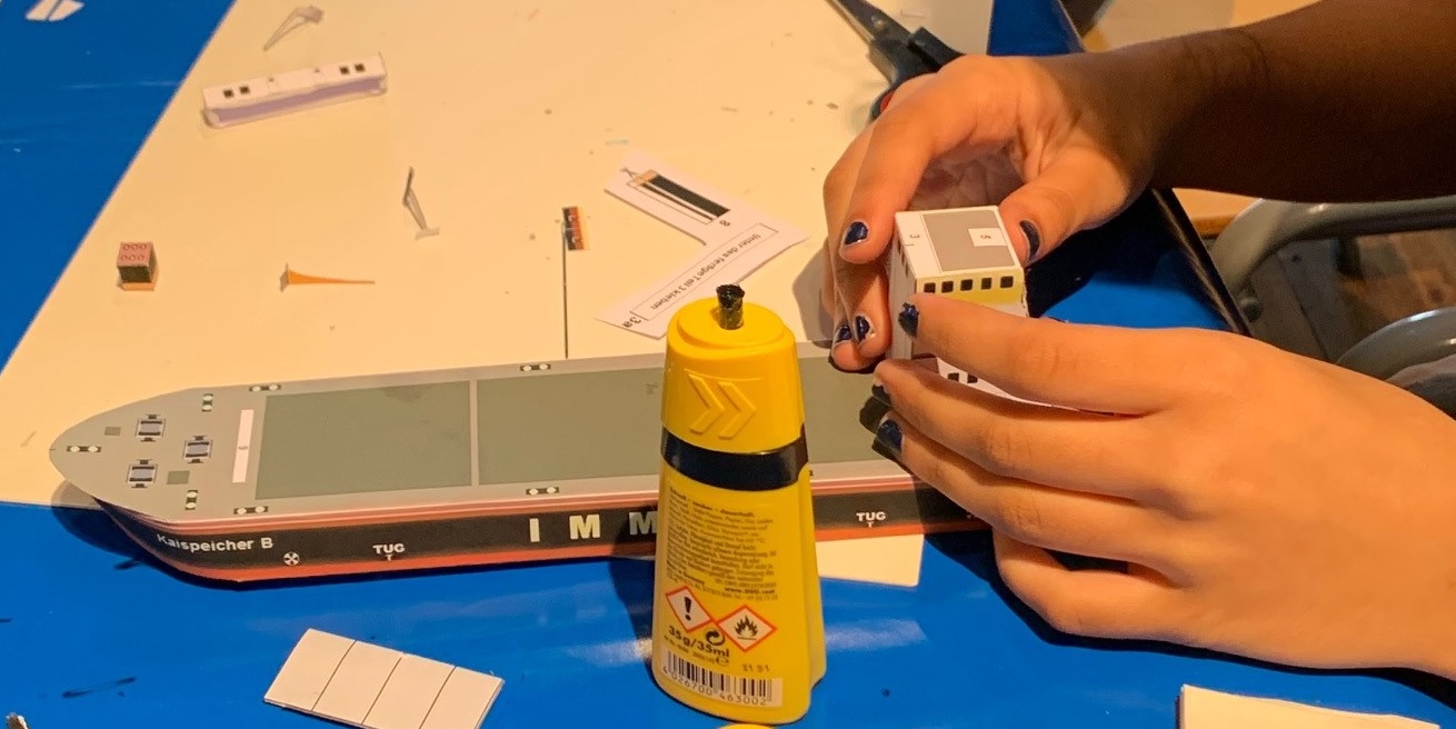 Zwei H&auml;nde mit dunklem Nagellack f&uuml;gen einen kleinen Papieraufbau an ein Papiermodellschiff, vor ihnen steht eine gelbe Klebstoffflasche auf einem blauen Tisch, umgeben von ausgeschnittenen Papierteilen und einer Schere.